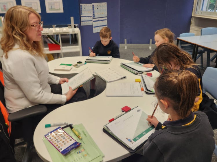 A teacher sitting at a jellybean table with 4 Stage 1 students, who are writing